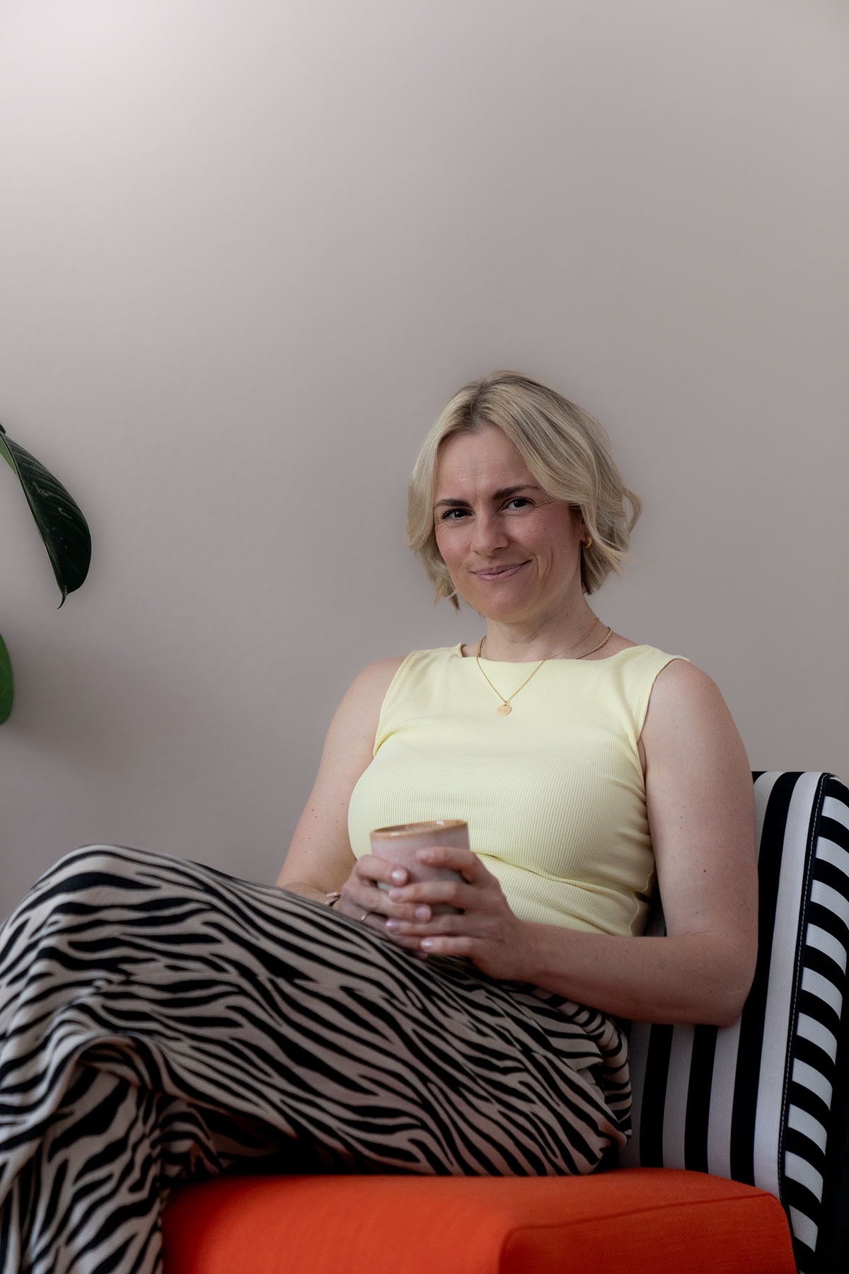 Judith Storm sitzt mit einer Tasse auf einer Bank mit gestreifter Lehne und lächelt in die Kamera.